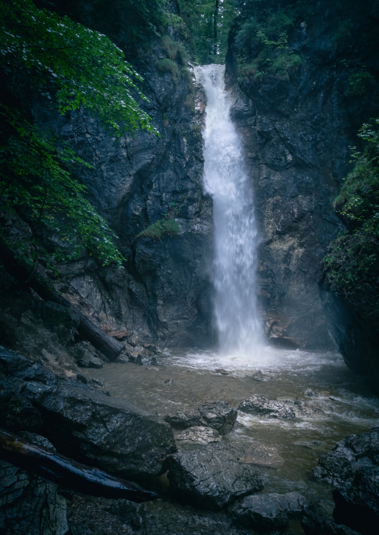 This image captures the mesmerizing beauty of a waterfall nestled deep within a forest. The waterfall, the central figure in this scene, gushes forth with great force. It courses down, contributing to a watercourse and possibly a mountain river, adding to the richness of water resources in the area. The waterfall is surrounded by lush, verdant trees, indicative of a nature reserve or state park. The landscape is teeming with life, with the possibility of a mineral spring, a creek, and a ravine in the vicinity. The terrain suggests that this could be a mountainous region with a valley nearby. The close-up details of the rocks and the trees add a raw, untouched feel to the image, enhancing its overall allure. Amidst the dense greenery, one can also spot traces of autumn with leaves beginning to change color, suggesting the onset of fall. This enchanting display of nature reaffirms the wonders of the outdoor world.